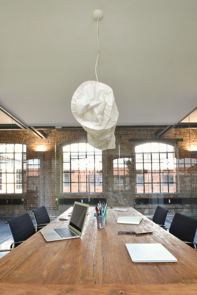 Wooden meeting table under a paper pendant lamp, with glass walls and factory windows behind it