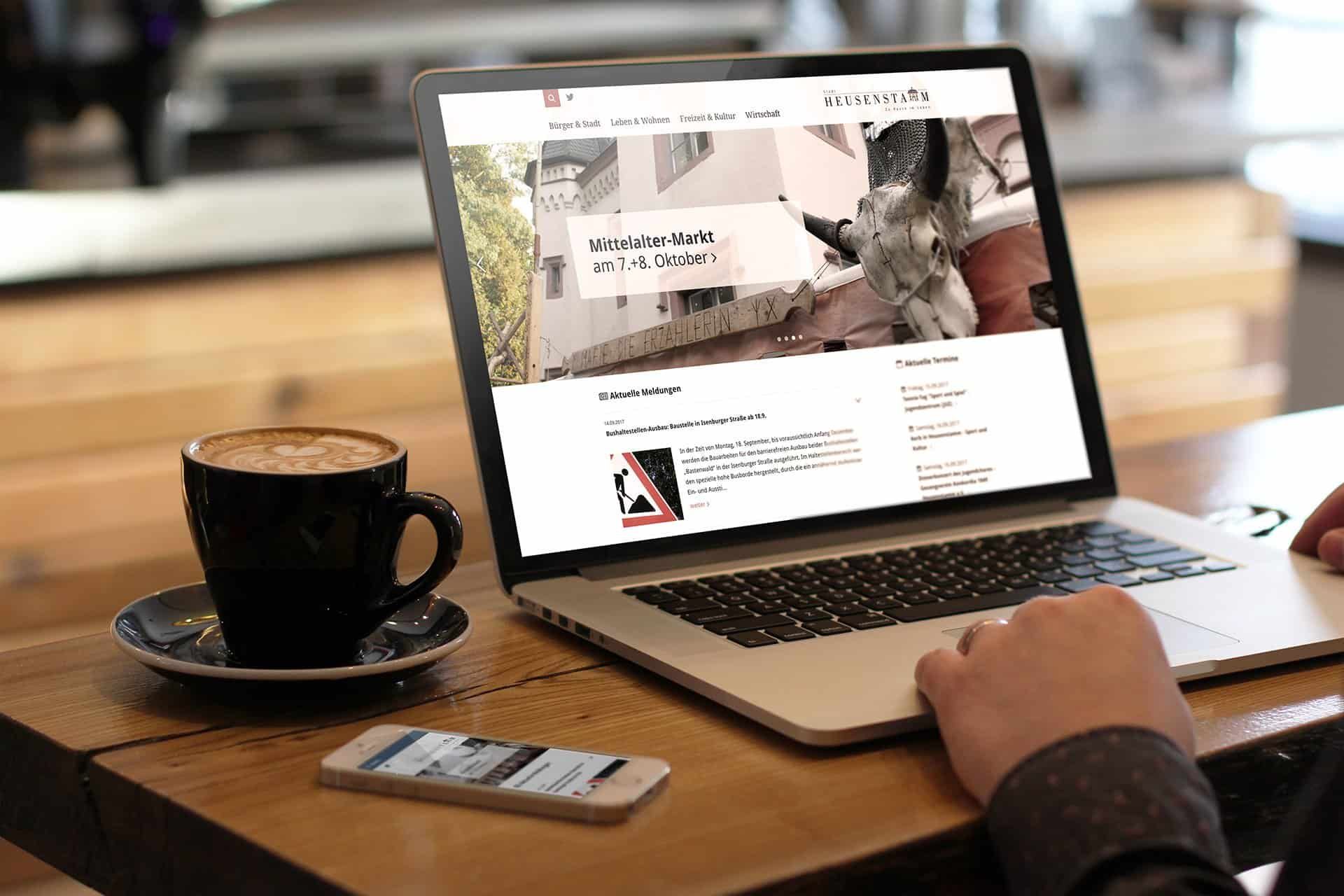 Laptop on a wooden table showing the heusenstamm.de home page, with a cappuccino and a smartphone beside it