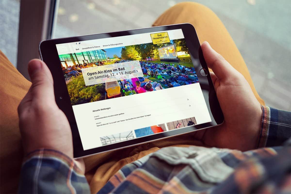 Tablet on a lap showing the Bad Heusenstamm pool website with a notice about an open-air cinema at the pool