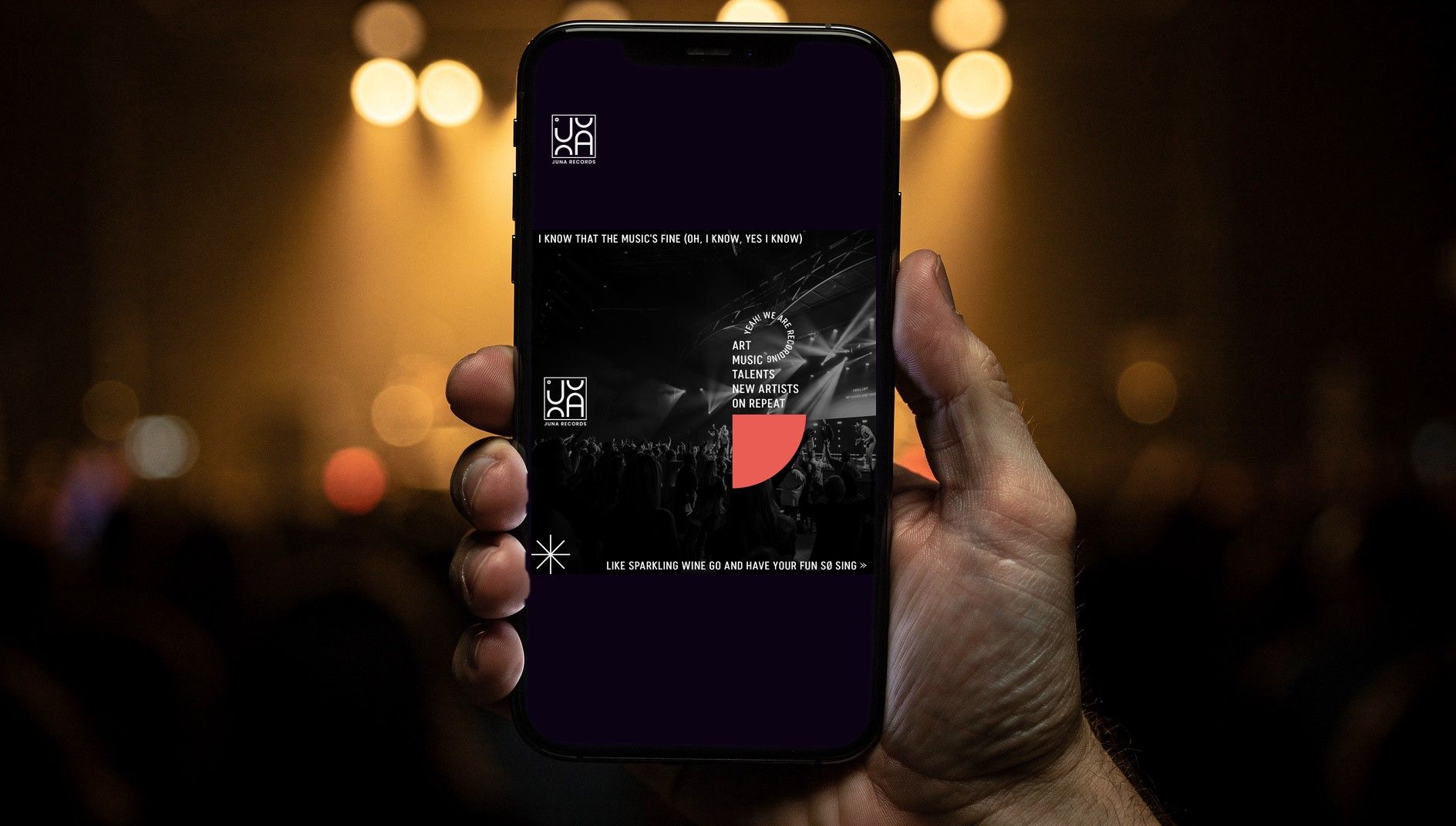 Hand holding a smartphone against stage lights, the screen showing a JUNA RECORDS social media post with a concert photo
