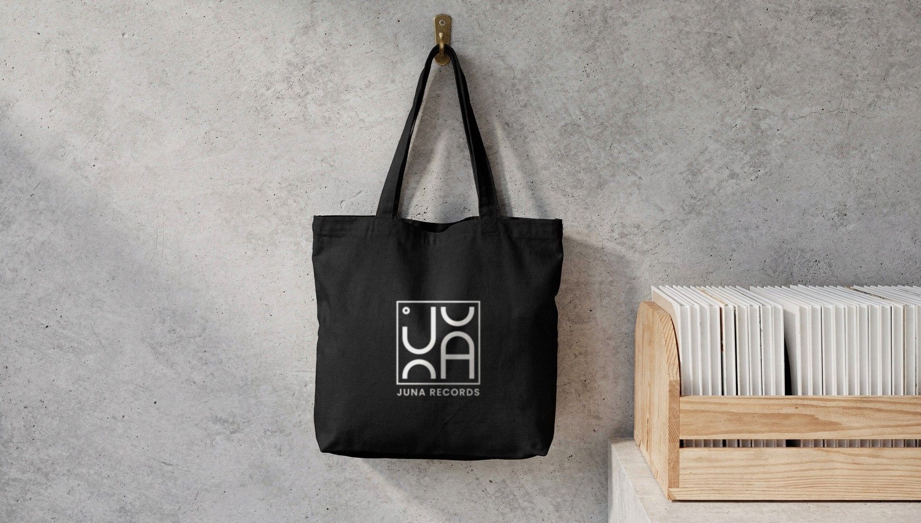 Black cotton tote bag with the white JUNA RECORDS logo hanging from a hook on a concrete wall