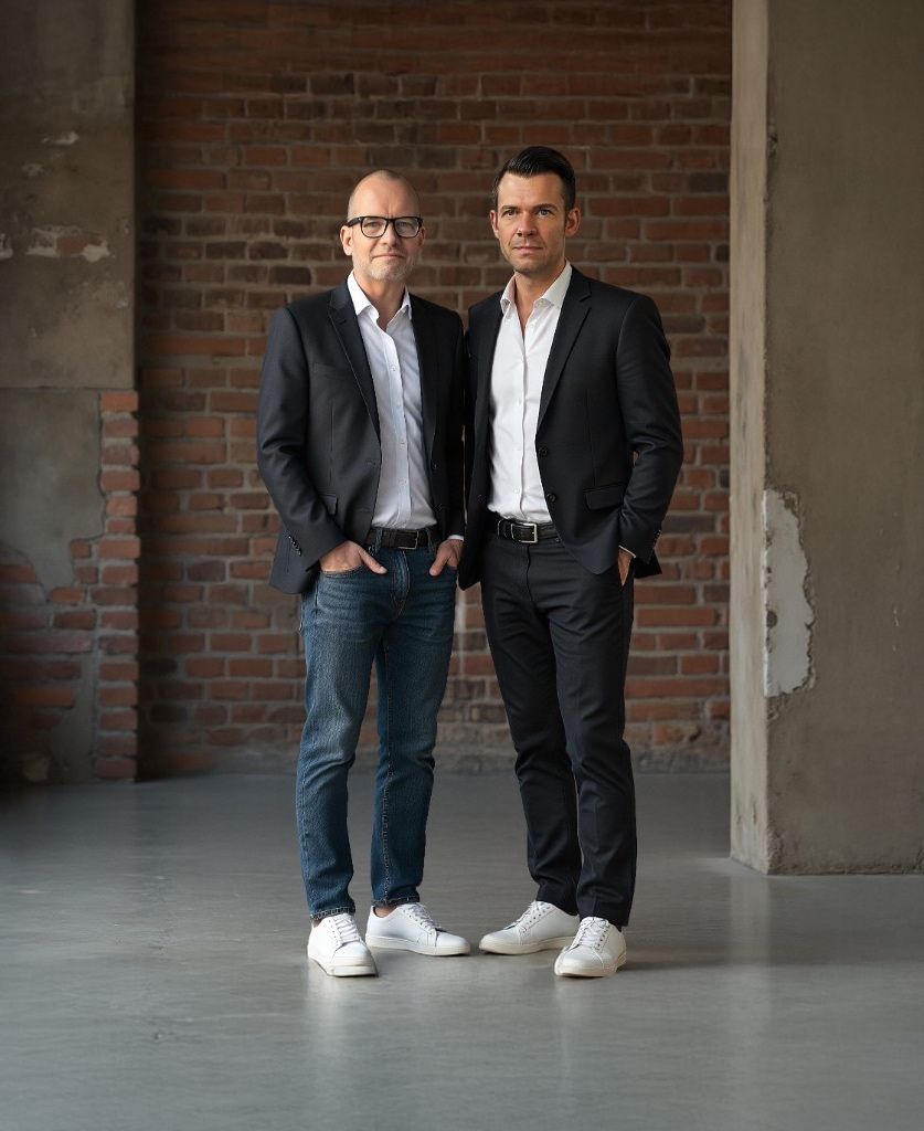Julian M. Flockton (left) and Markus Zebisch (right) in blazers and white trainers in front of a brick wall