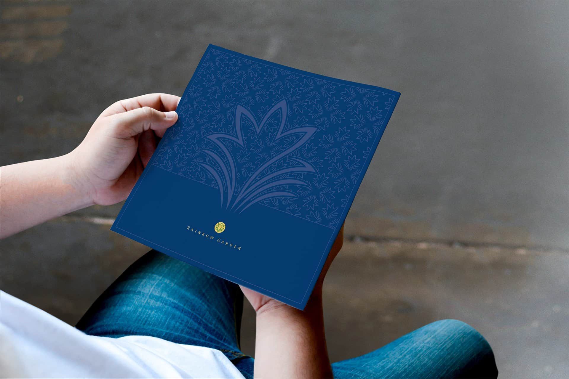 Hands holding a blue Rainbow Garden brochure with a flower pattern and a gold logo
