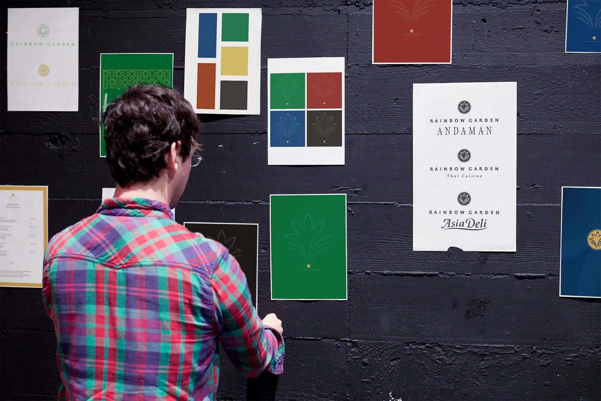 A man putting up posters on a dark wall: colour charts, patterns and the logos of Rainbow Garden Andaman, Thai Cuisine and Asia Deli